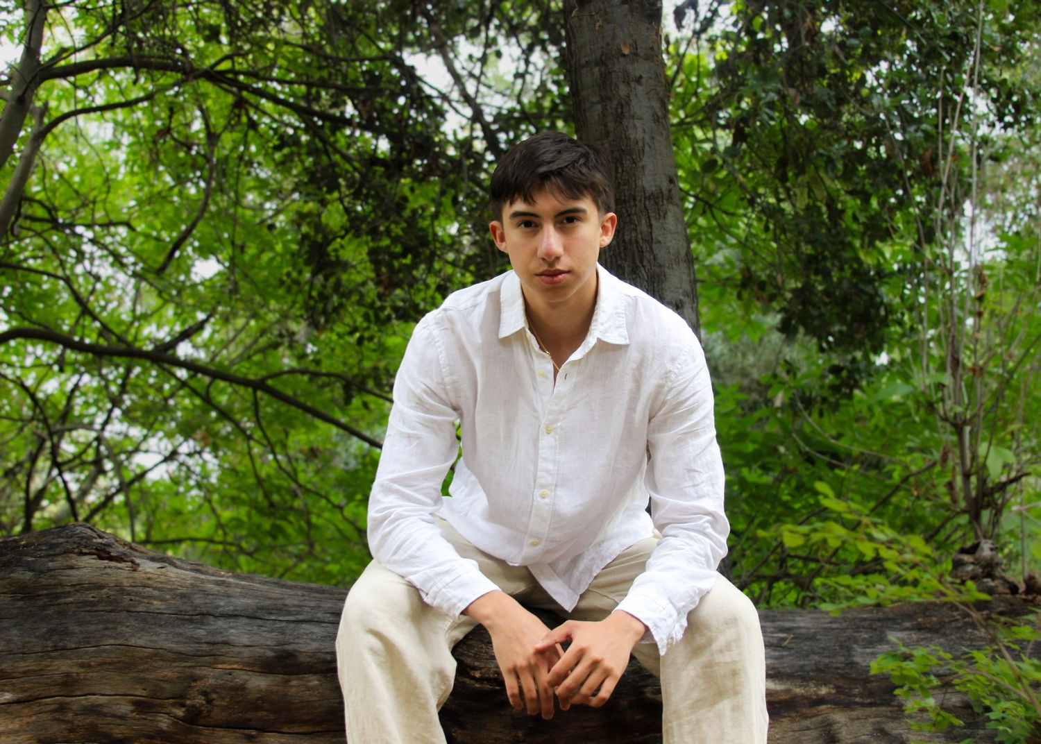 Young man in white shirt and khaki pants sitting on a fallen log surrounded by lush green trees.