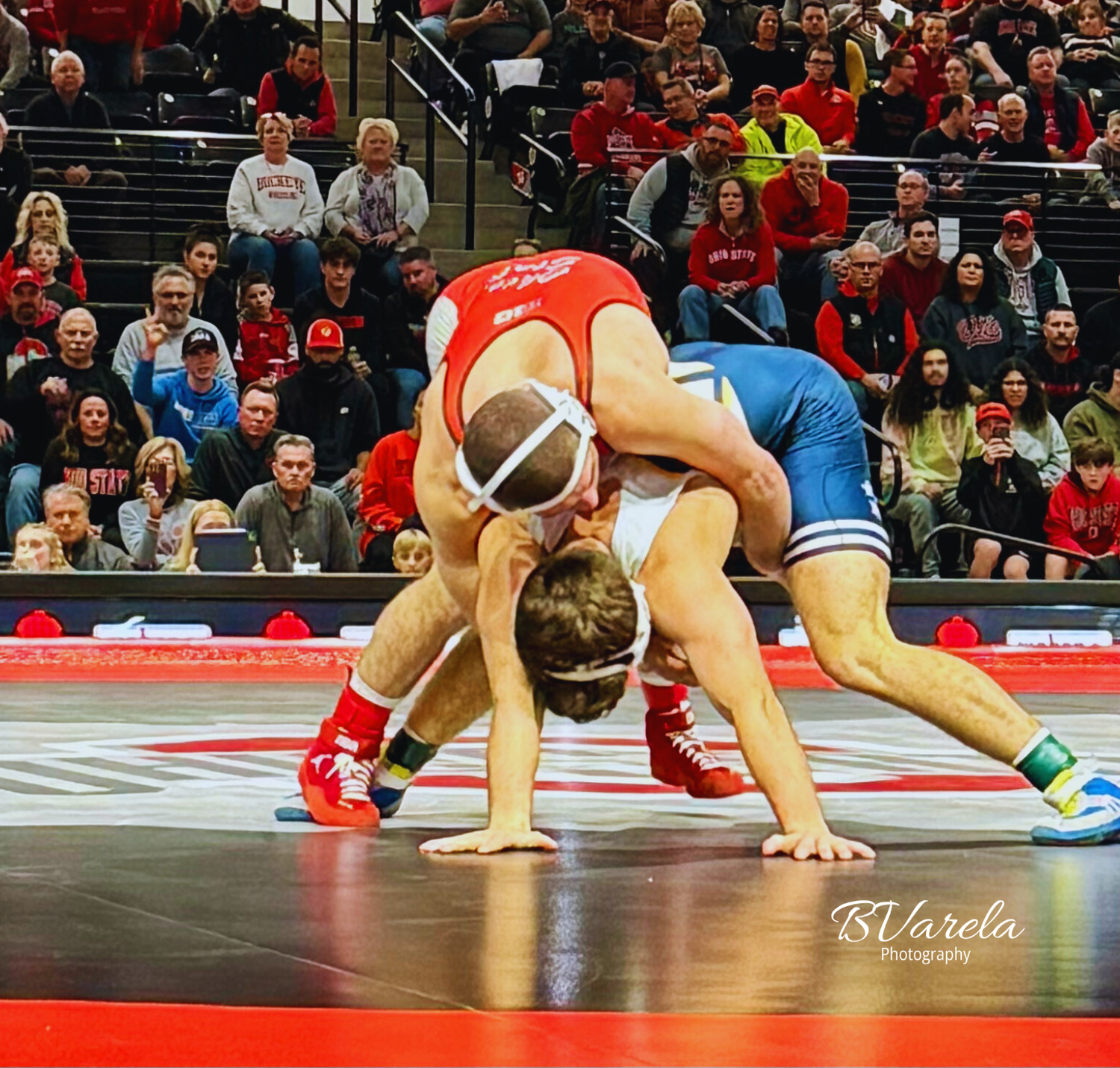 Two wrestlers compete on a red mat at a college wrestling match with a packed crowd in the background.