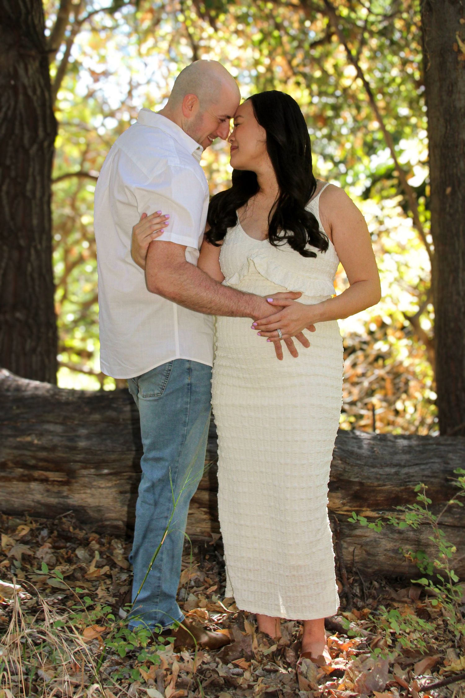 Couple sharing a tender moment outdoors, pregnant woman in white dress with partner in wooded setting.