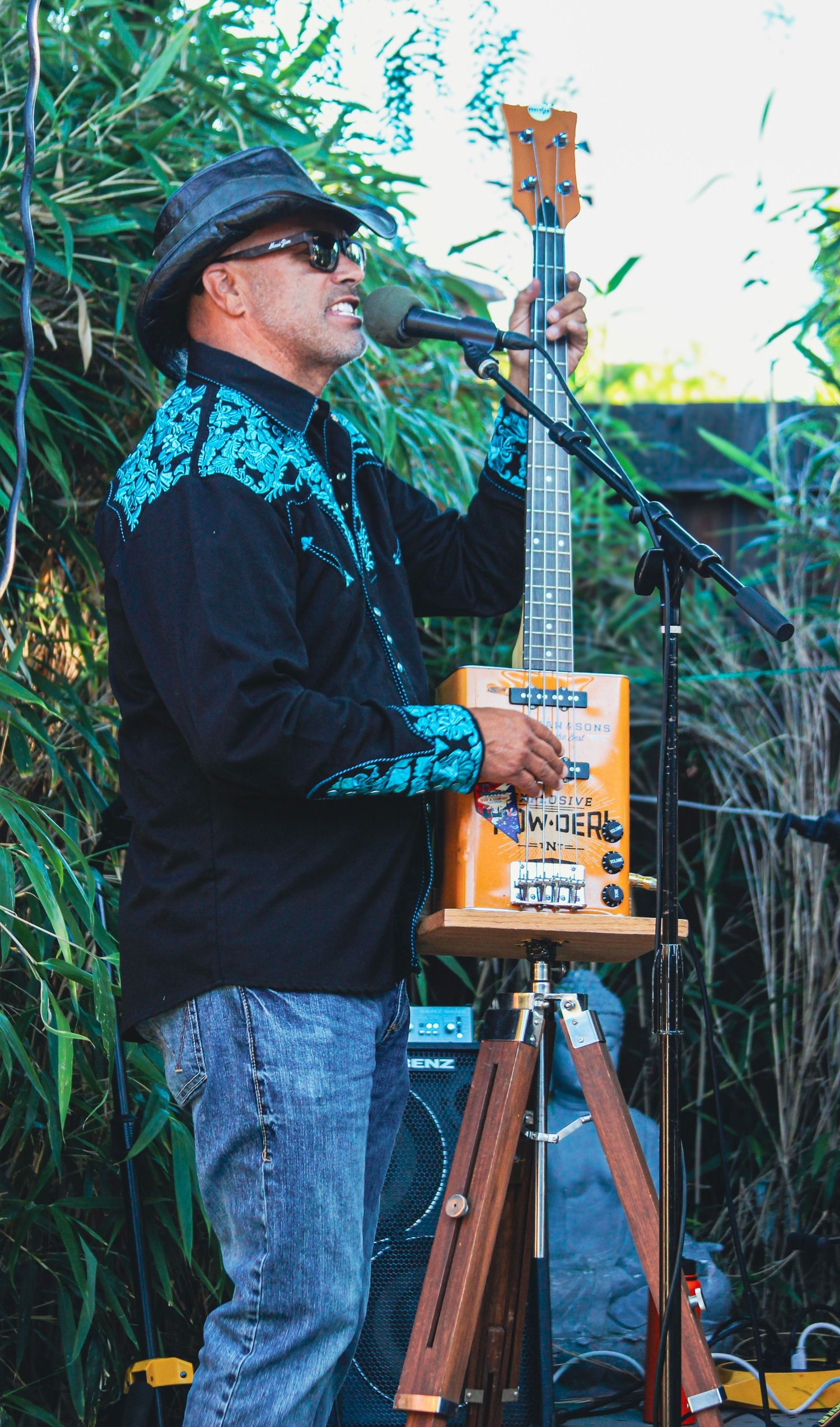 Man in black shirt and hat plays cigar box guitar at outdoor microphone stand surrounded by lush green foliage.
