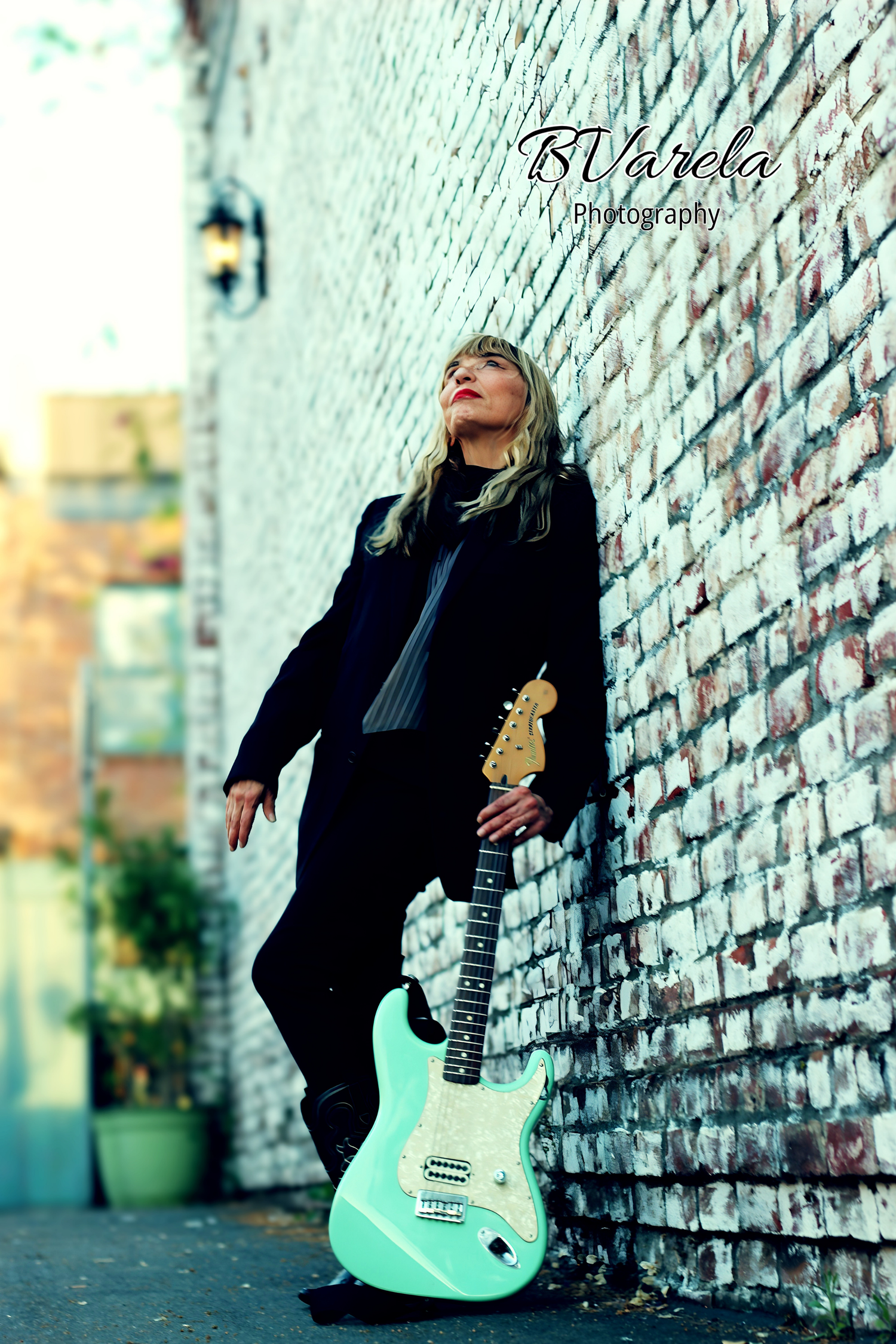 Woman in black outfit leaning against brick wall holding a seafoam green Fender Stratocaster guitar outdoors.