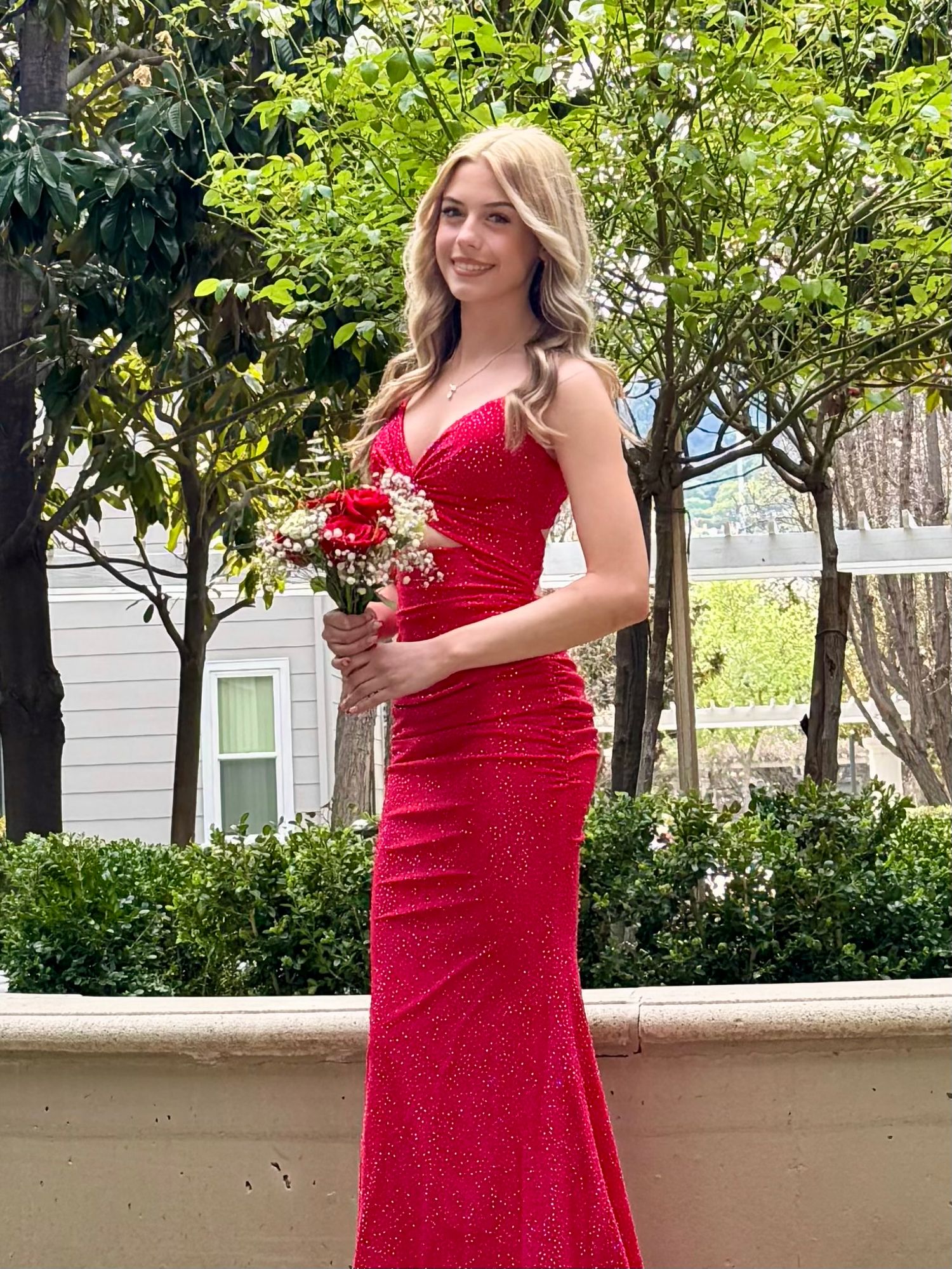 Young woman in a sparkling red formal gown holds a red bouquet, posing outdoors for prom.