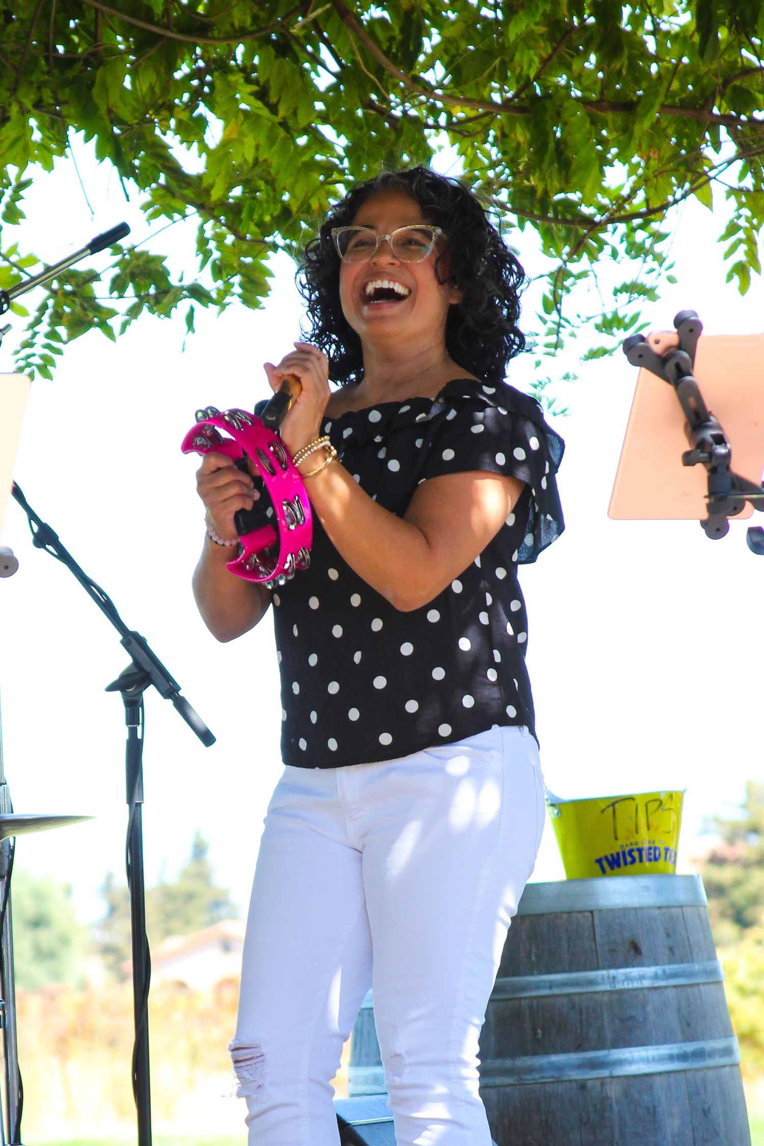 Woman in polka dot top and white jeans laughs while playing pink tambourine on outdoor stage.