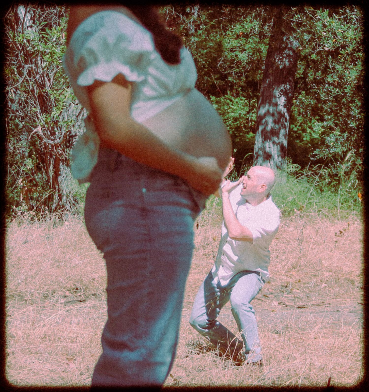 Pregnant woman standing outdoors near trees while a man crouches in the background during a maternity photo shoot.