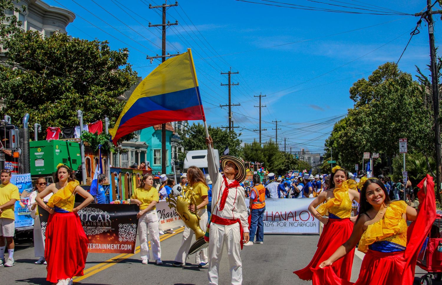 Colorful Colombian cultural parade with flag bearers and dancers in traditional yellow and red costumes on a sunny street.