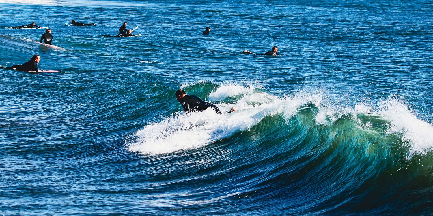 Surfers riding blue ocean waves in wetsuits, with a group waiting in the lineup on a sunny day.