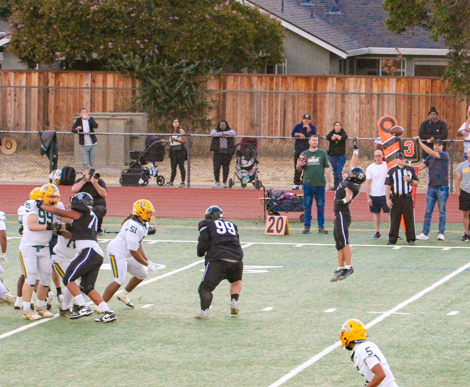 Youth football game in progress on a track field with players in yellow and dark uniforms, referees, and spectators watching.