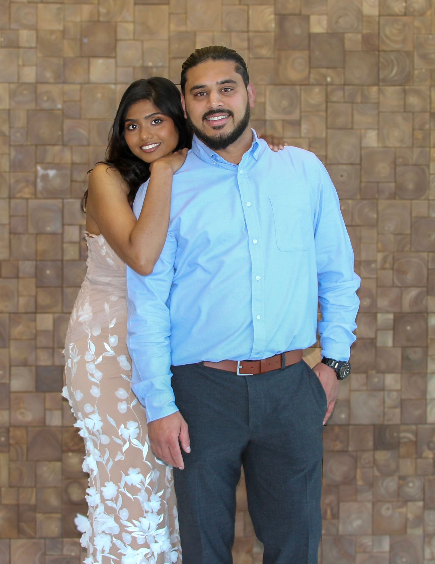 A smiling couple poses together, woman in floral gown and man in blue shirt, against a stone tile wall.