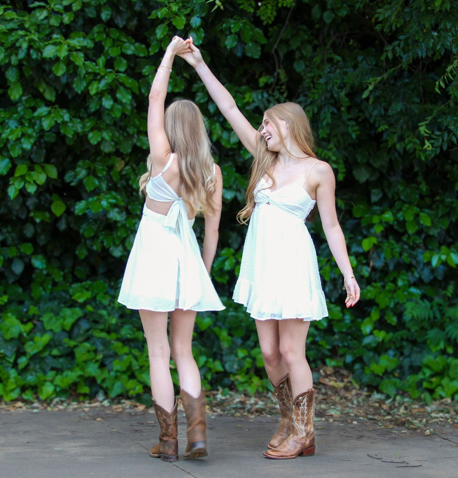 Two young women in matching white dresses and cowboy boots dancing together outdoors near a green hedge.