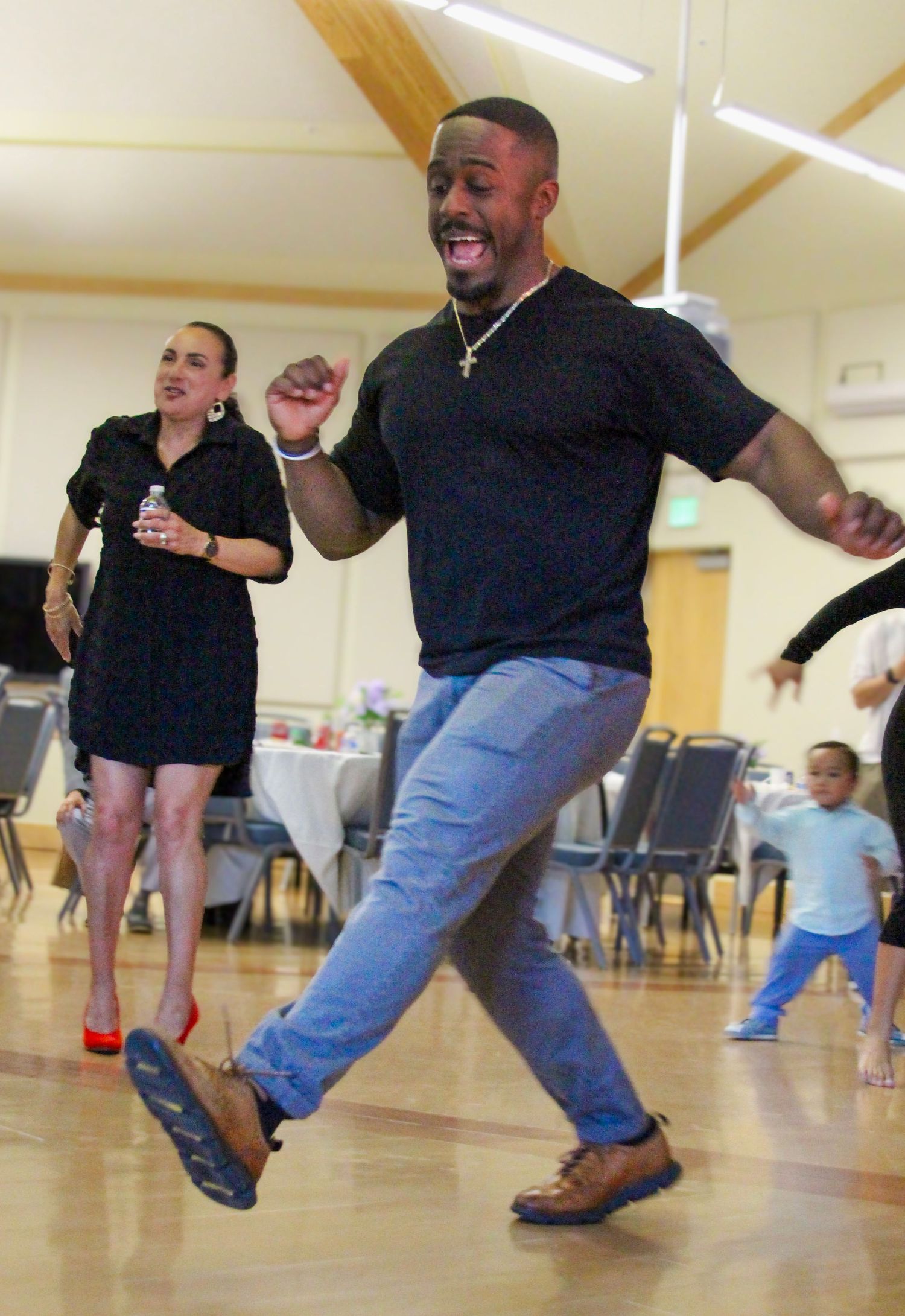 Man in black shirt and jeans dancing energetically at an indoor social event with others in the background.