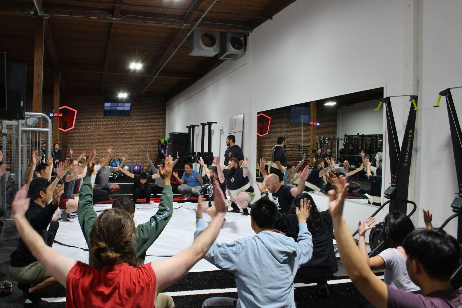 Large group fitness class with hands raised in a modern gym with mirrored walls and exercise equipment.