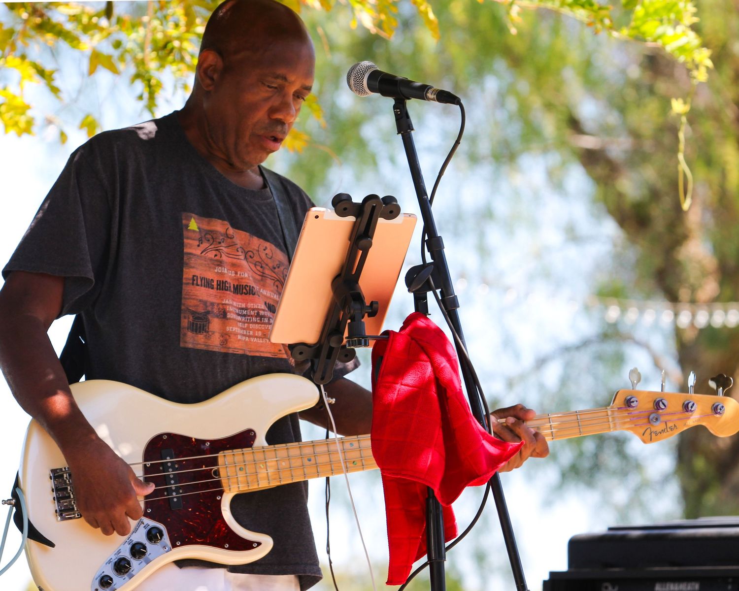 Man playing white bass guitar at outdoor concert with microphone stand and red cloth nearby.