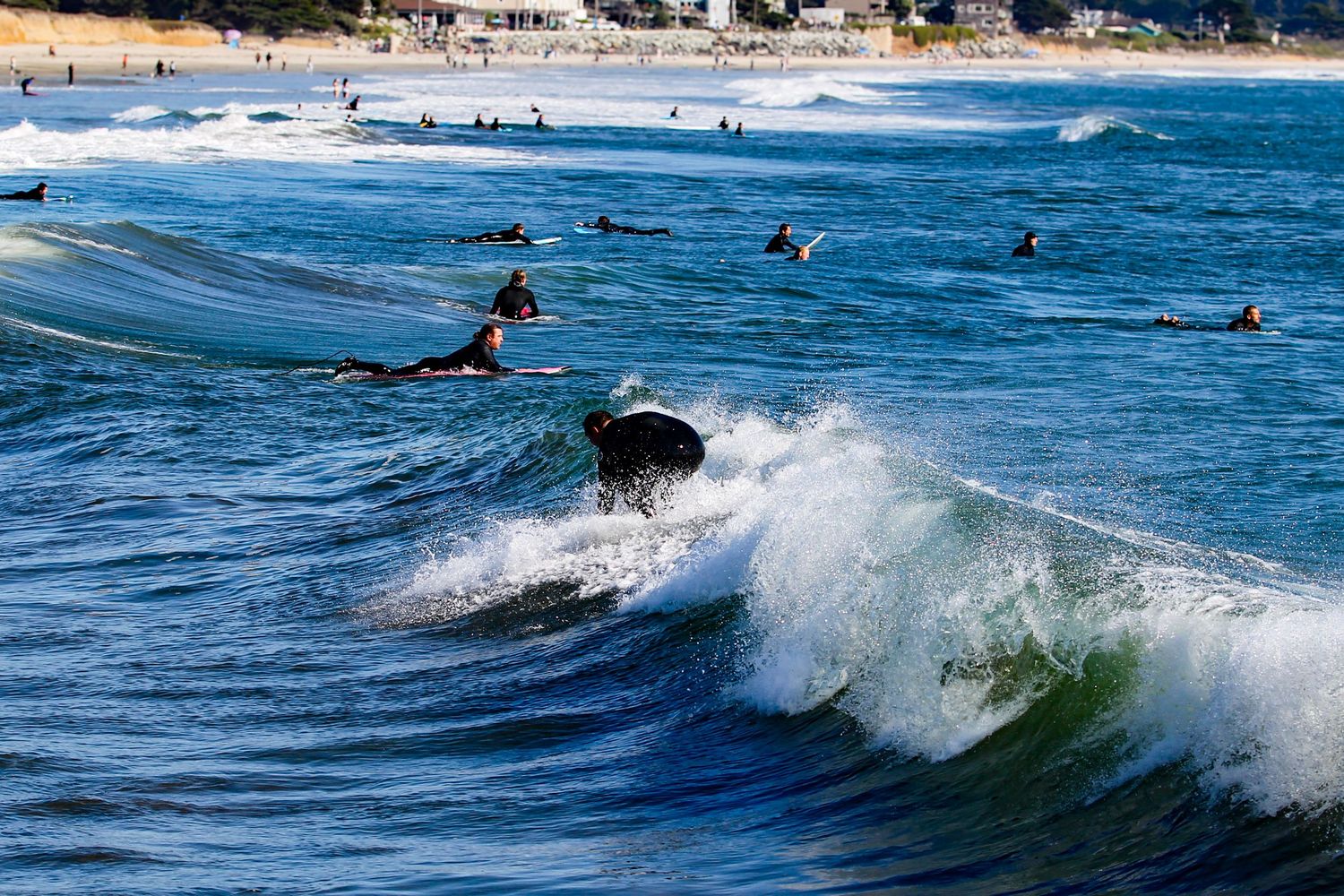 Surfers riding ocean waves at a sunny beach with many swimmers visible in the blue water and sandy shore in background.
