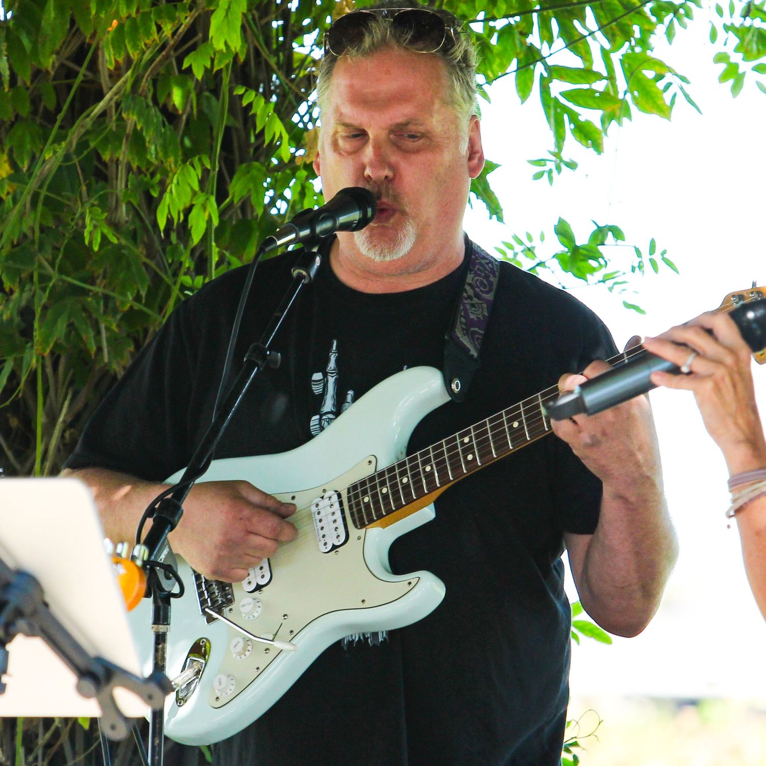 Man in black t-shirt playing white electric guitar and singing into microphone outdoors under green trees.