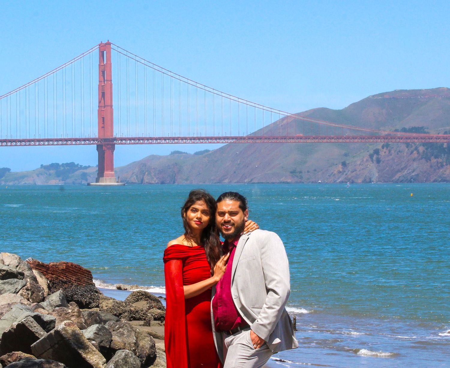 Couple posing at San Francisco's Golden Gate Bridge beach on a sunny day, woman in red dress, man in white suit.