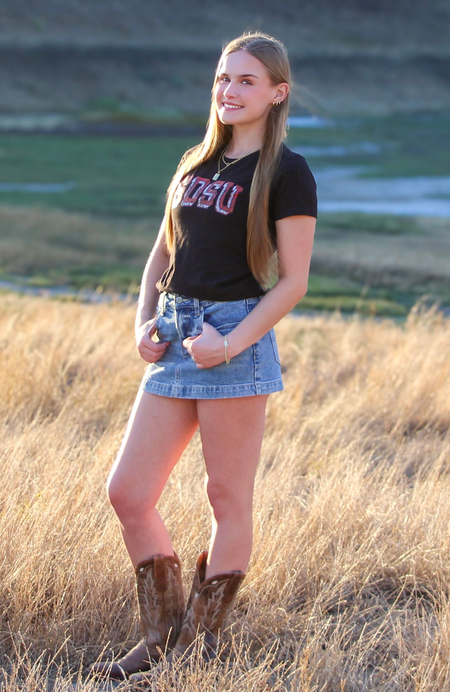 Young woman in SDSU shirt, denim skirt, and cowboy boots standing in a golden grass field.