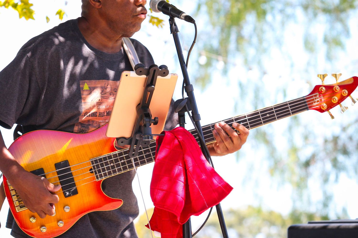 Male bassist plays colorful electric bass guitar on outdoor stage with microphone stand at live concert.