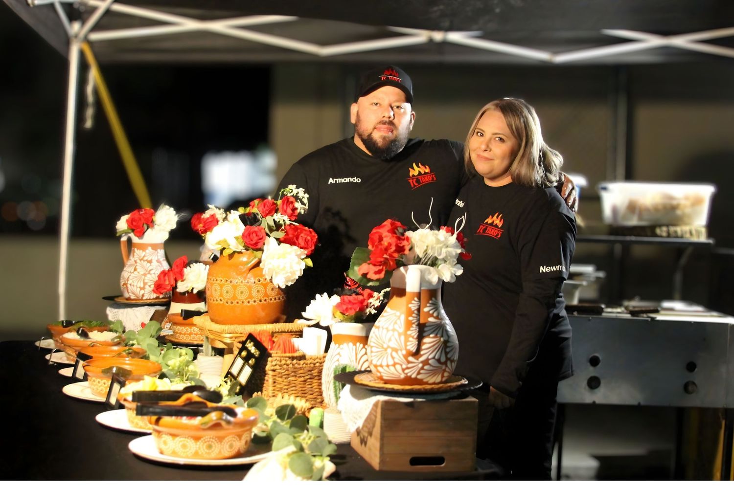 Two chefs stand behind an elegant food display table with floral arrangements and baked goods at a catering event.
