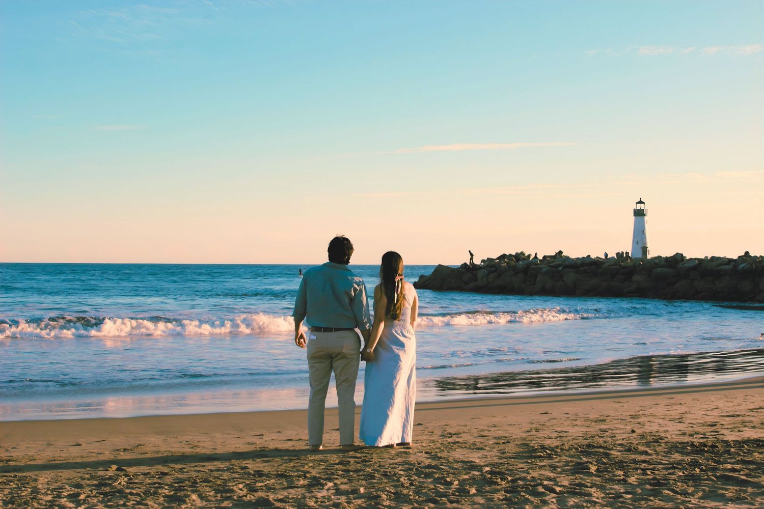 Couple holding hands on a sandy beach at sunset with a lighthouse in the background.