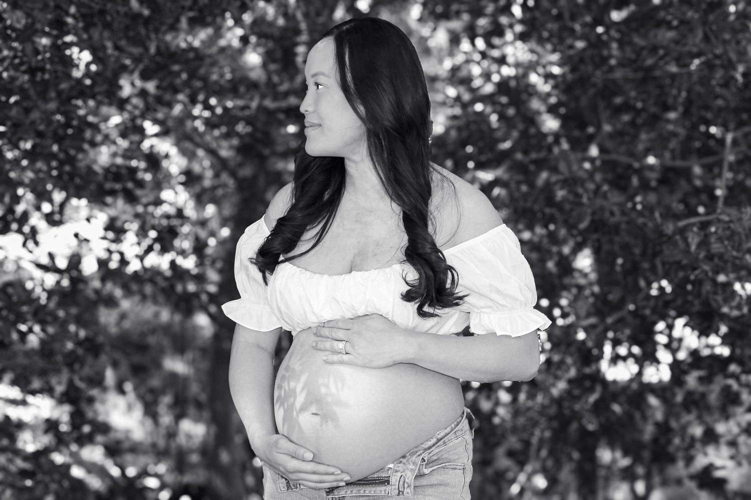 Black and white maternity photo of a pregnant woman in white off-shoulder top and jeans, cradling her baby bump outdoors.