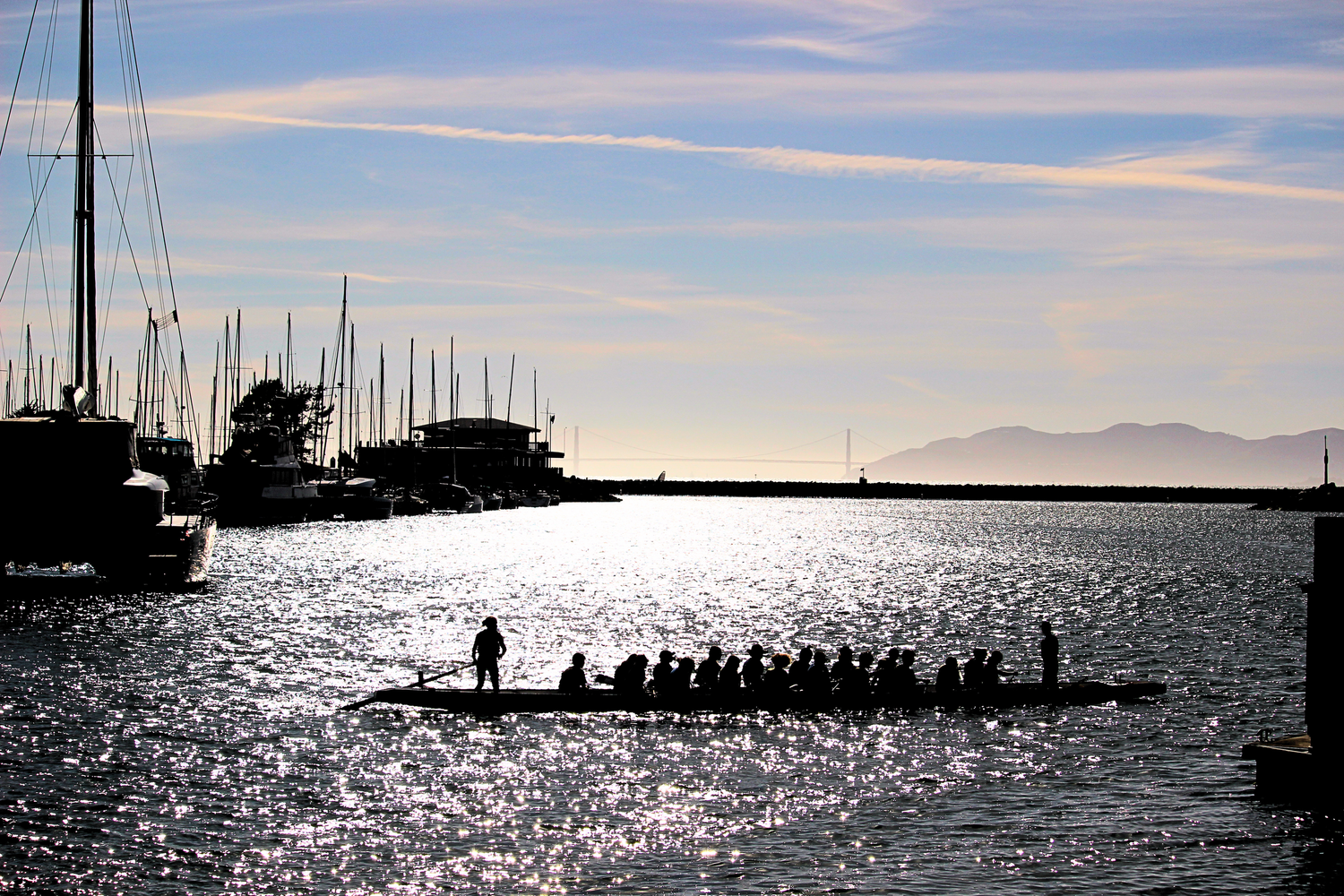 Silhouette of a dragon boat team rowing at sunset near a marina with sailboats and mountains in the background.