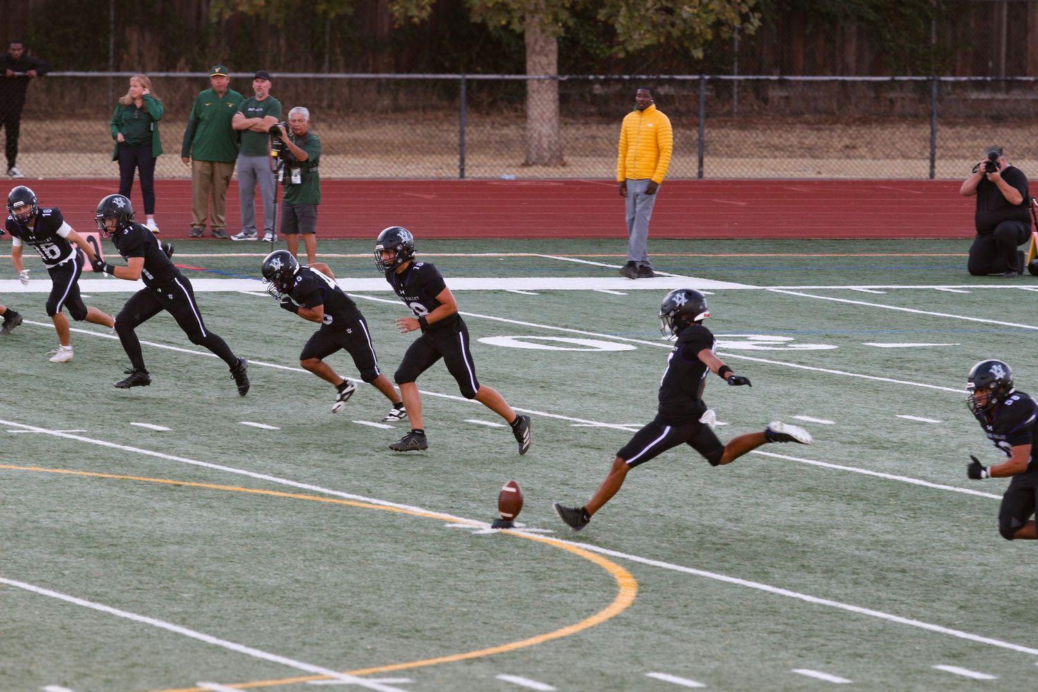 Youth football players in black uniforms line up on a green turf field during a kickoff play.