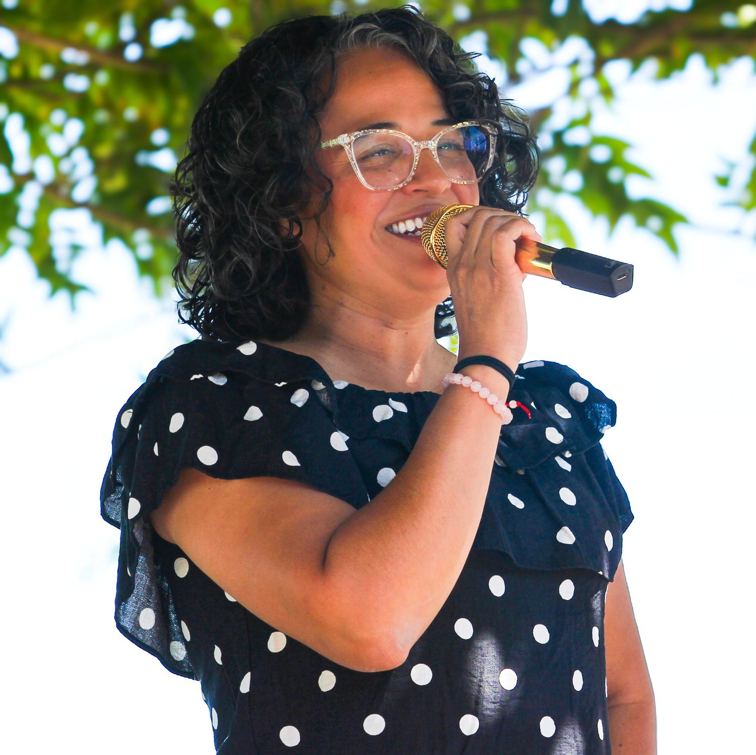 Woman in polka dot dress singing into microphone outdoors, wearing glasses at a live performance event.