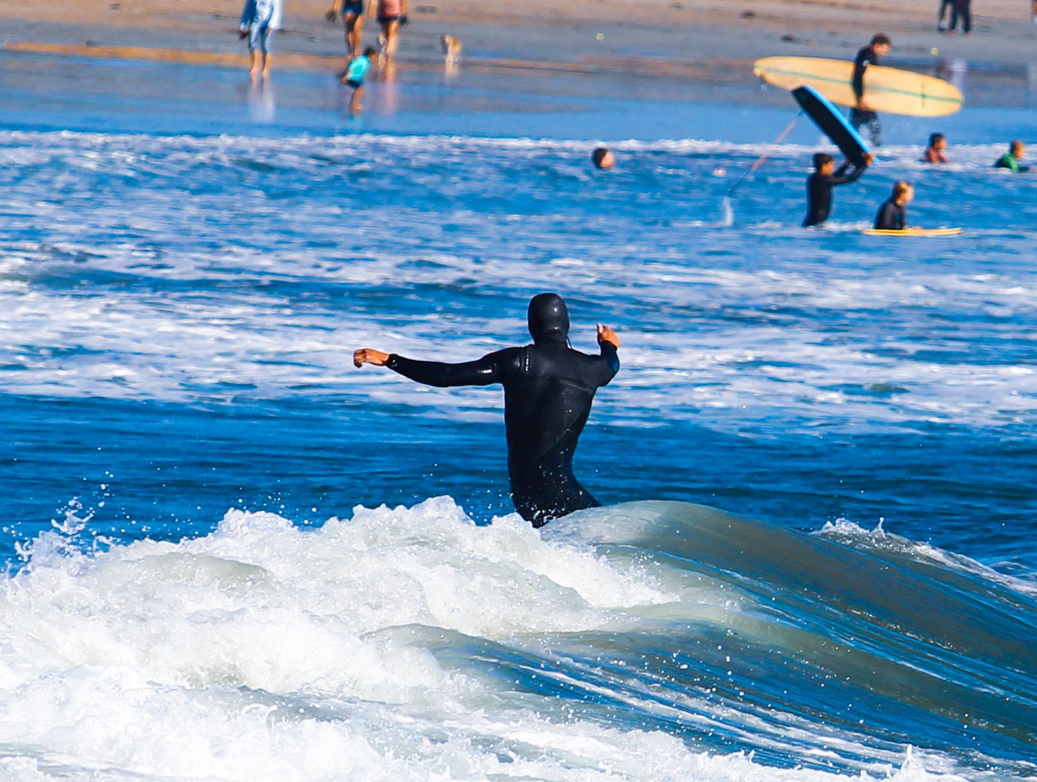 Surfer in black wetsuit riding waves at a busy beach with other surfers visible in the background.
