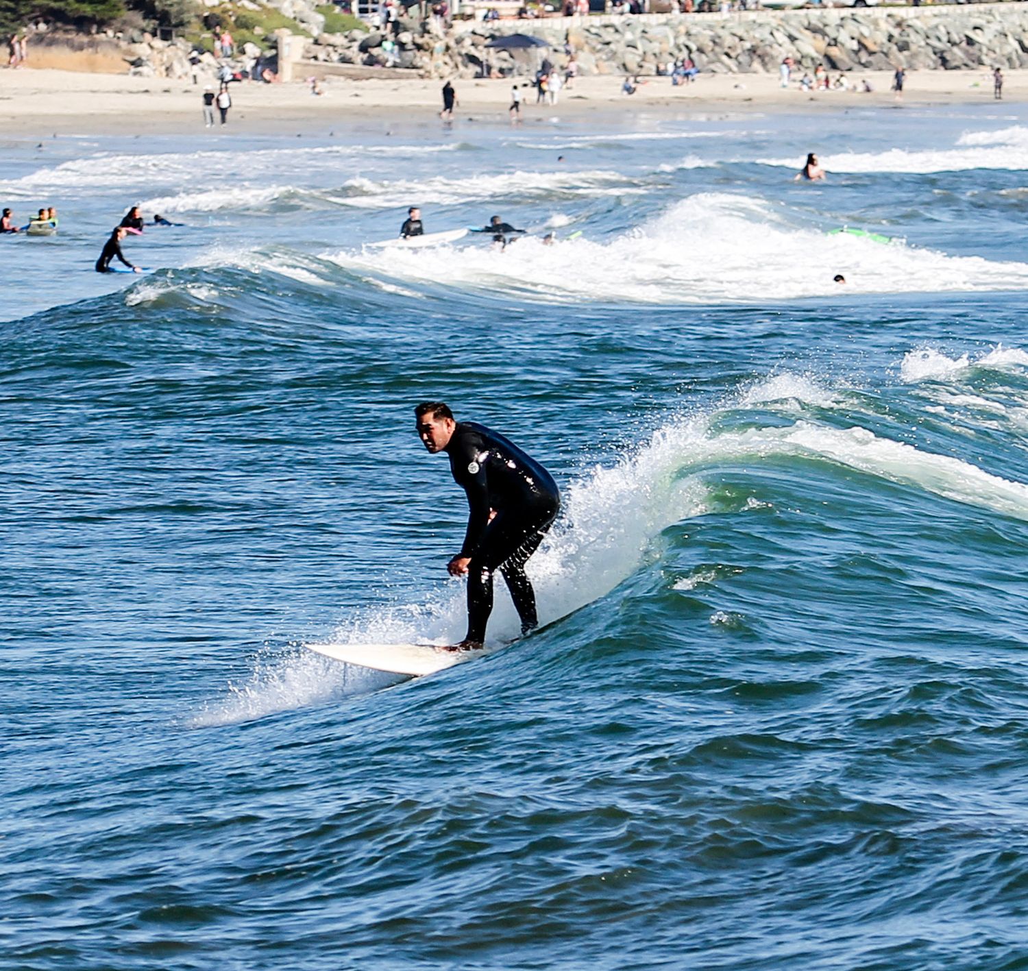 A surfer in a black wetsuit rides a wave at a crowded beach with other surfers visible in the background.