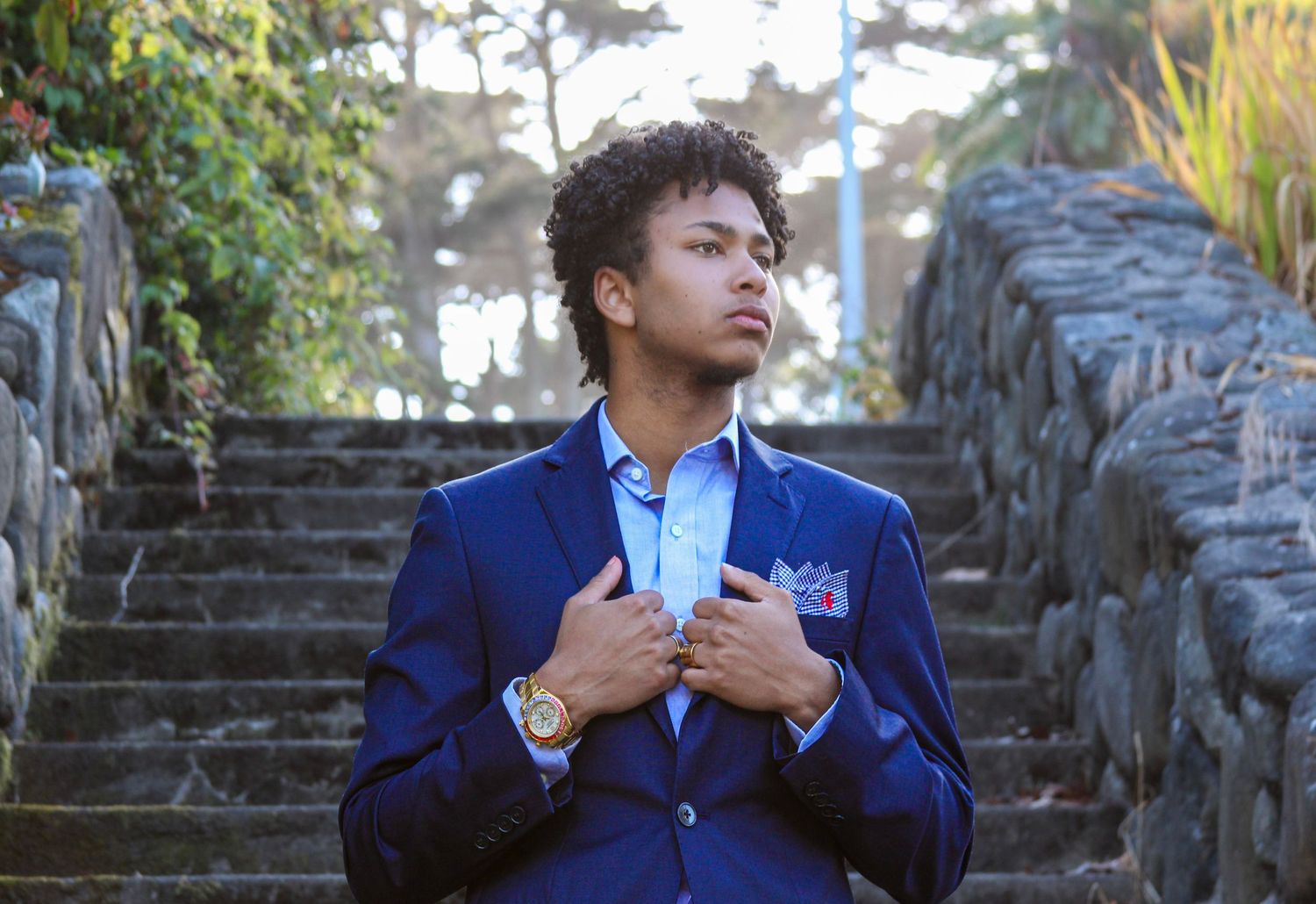 Young man in a navy blue suit with a pocket square poses confidently on stone steps outdoors.