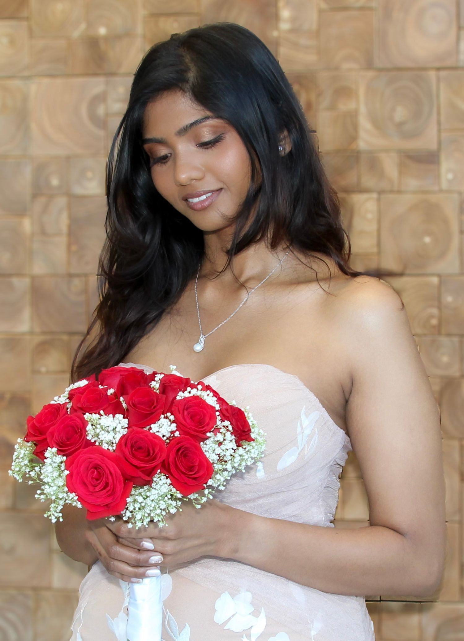 Young woman in strapless gown holding red rose bouquet with baby's breath, smiling elegantly at a formal event.