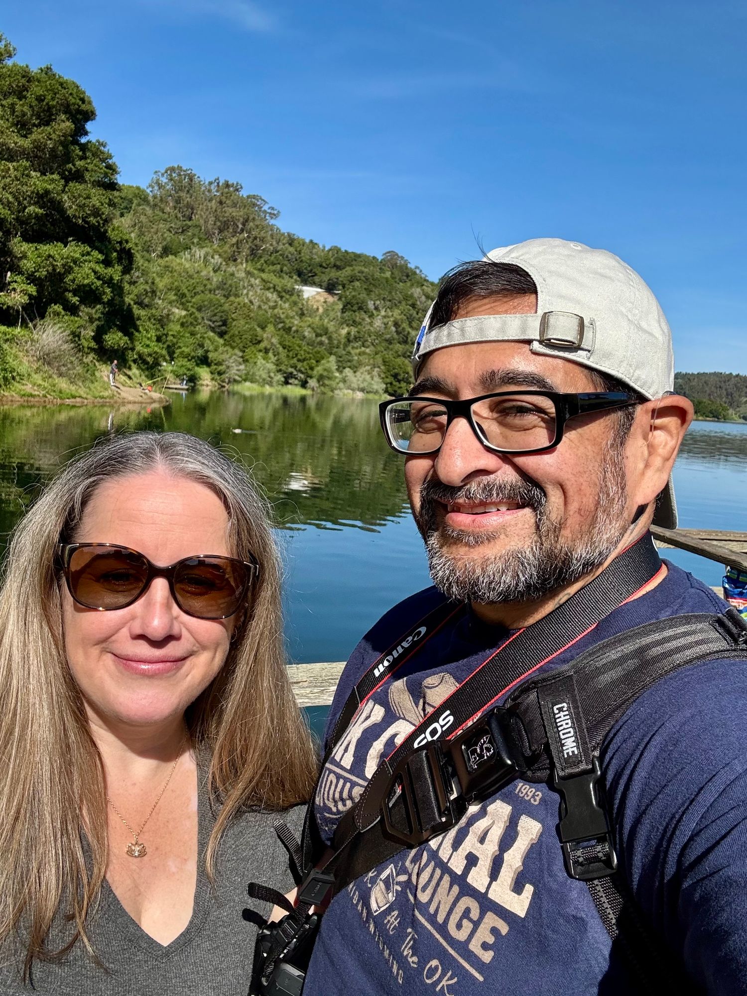 Two people smiling for a selfie outdoors near a calm lake surrounded by lush green trees under a clear blue sky.