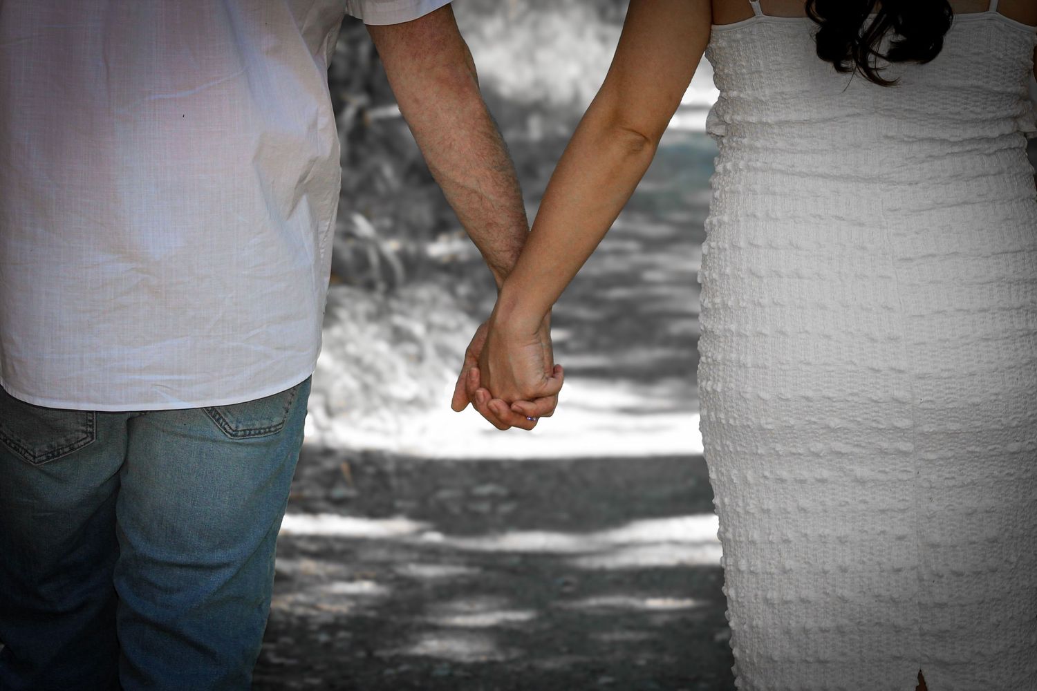 Couple holding hands while walking on a shaded path, man in jeans and white shirt, woman in white dress.