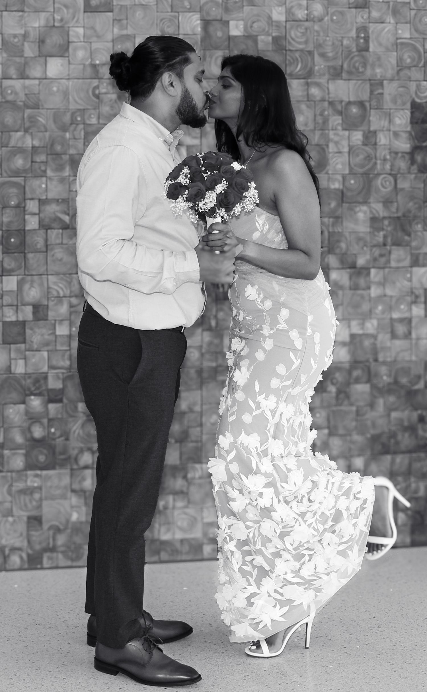 Black and white photo of a couple kissing at their wedding, bride in floral gown holding bouquet, groom in white shirt.