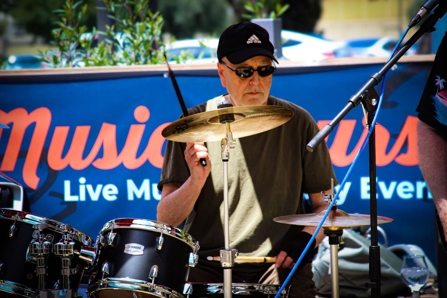 Musician playing drums at an outdoor live music event, wearing sunglasses and a black cap.