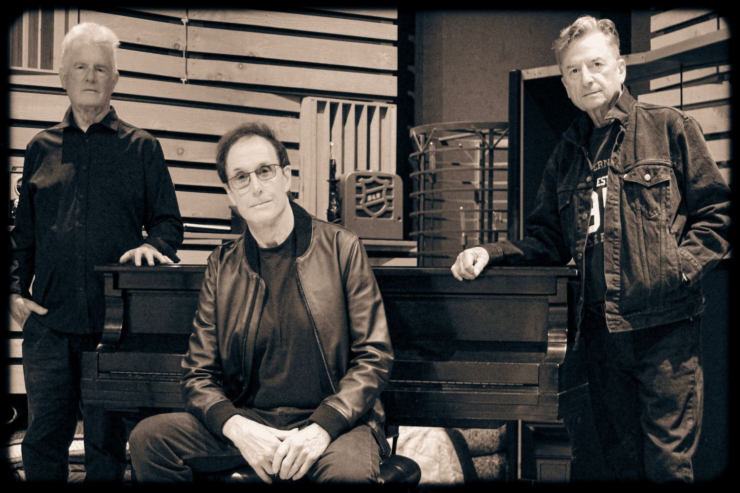 Three men pose in a recording studio in a black and white photo, two standing and one seated at a piano.