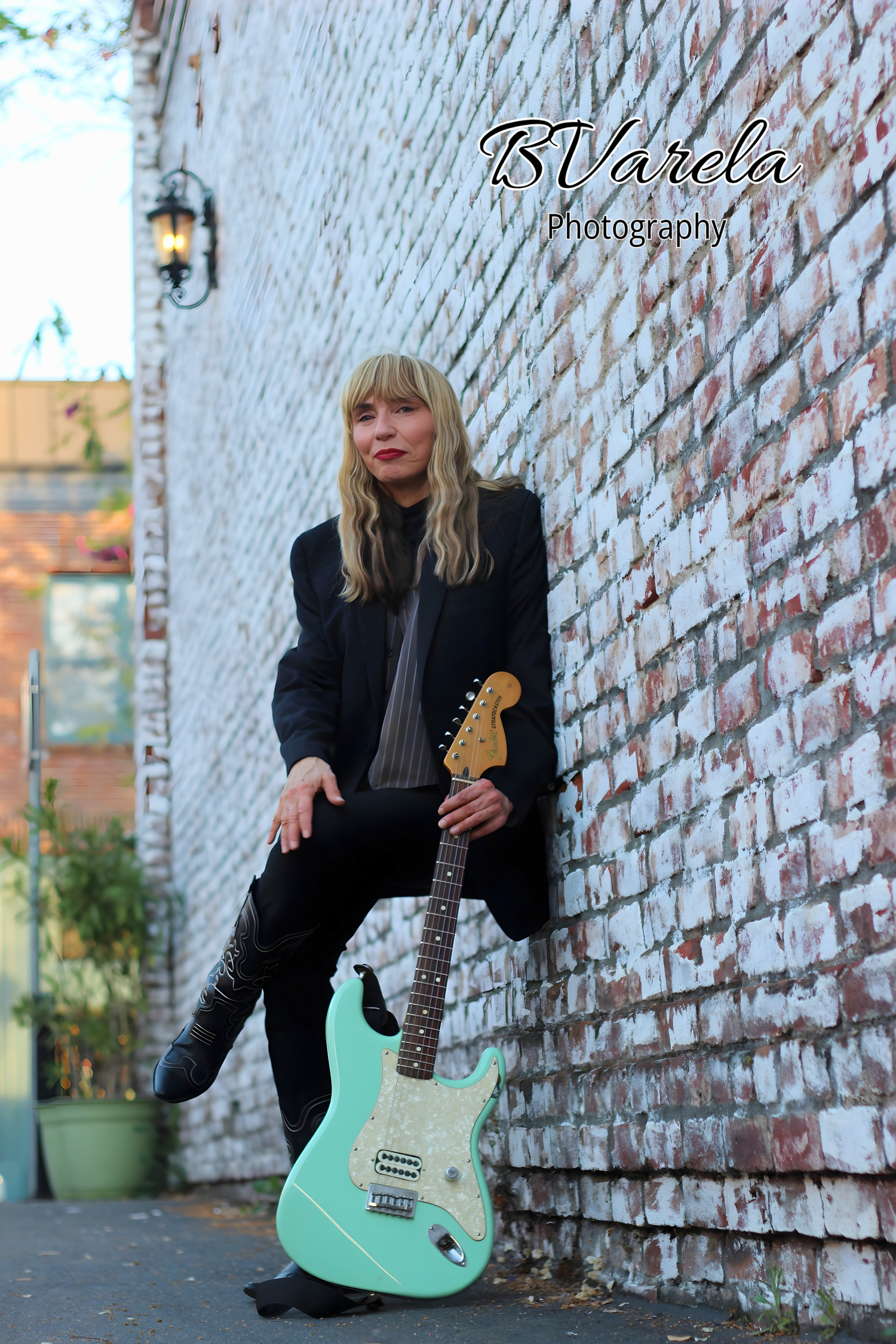 Blonde woman in black jacket poses with mint green Fender Stratocaster guitar against rustic brick wall outdoors.