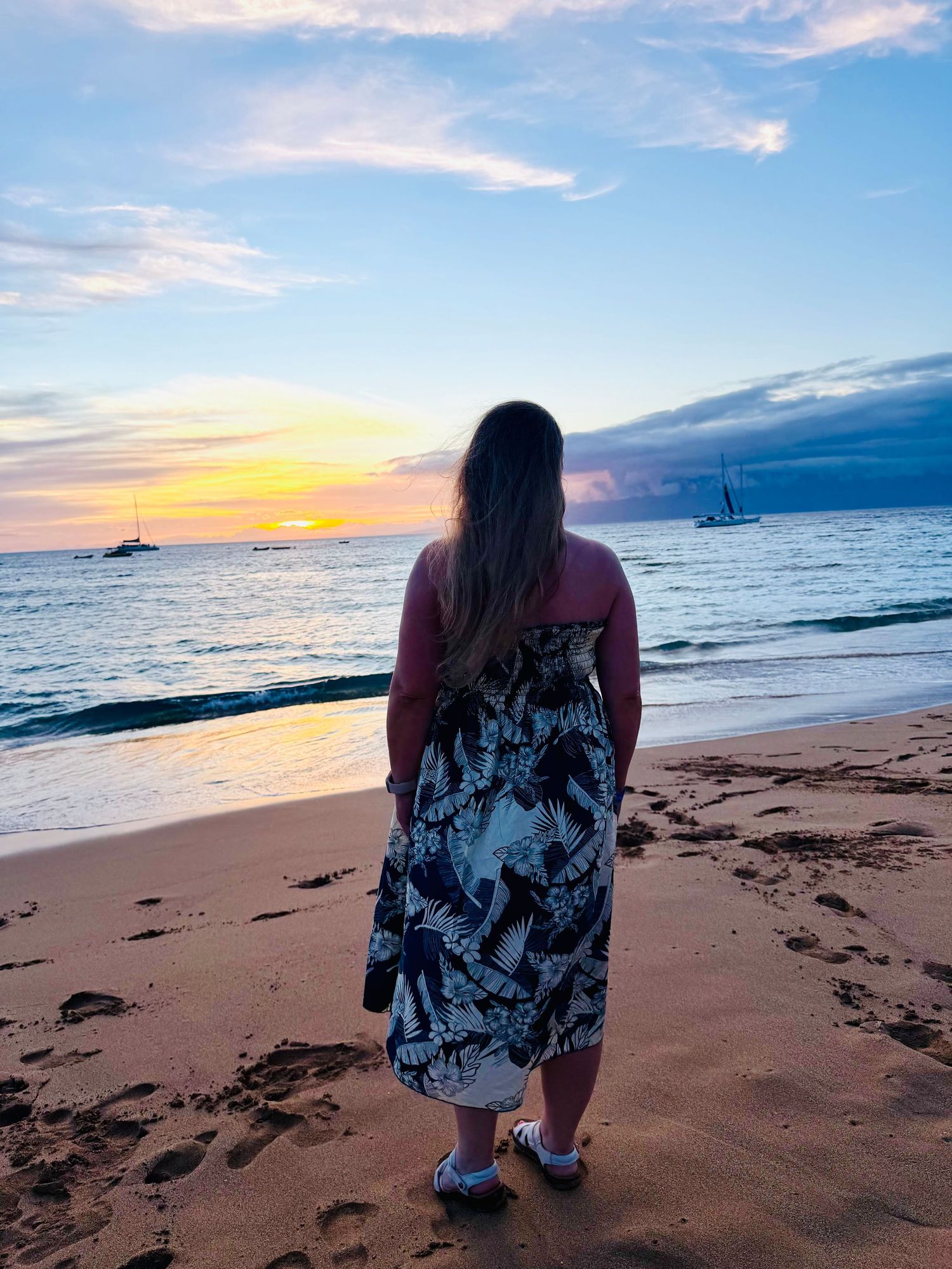 Woman in floral dress standing on sandy beach watching vibrant sunset over calm ocean with sailboats in distance.