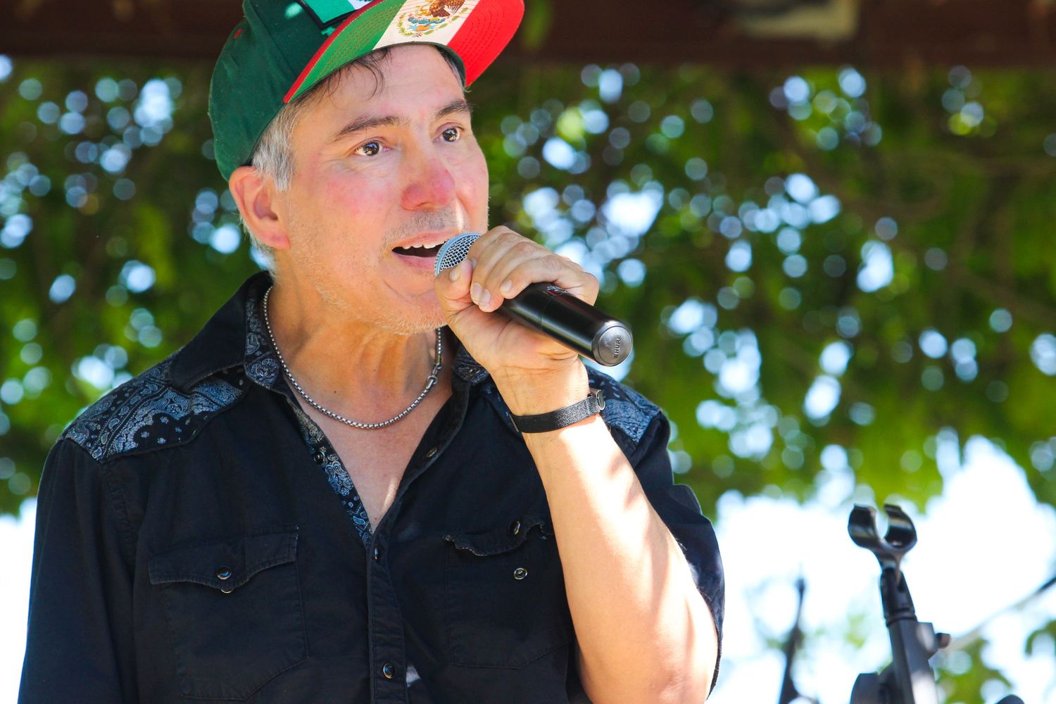 Middle-aged man singing into microphone outdoors, wearing black shirt and green Mexican flag cap at live event.
