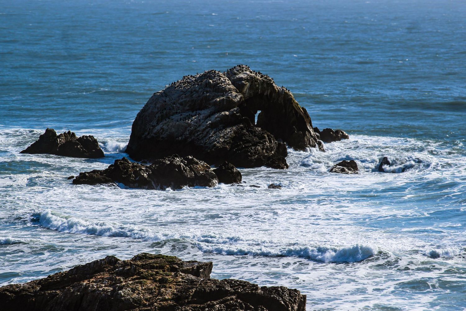 Rocky coastal outcropping surrounded by crashing ocean waves under a clear blue sky.