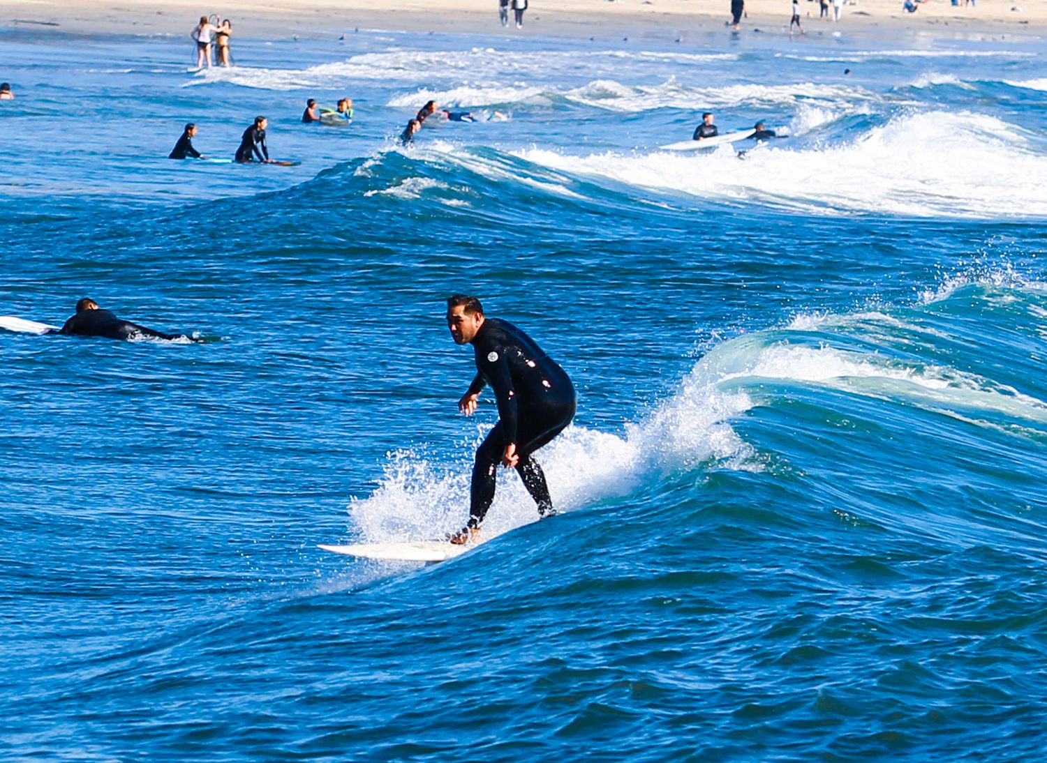 A surfer in a black wetsuit rides a wave at a busy beach with other surfers visible in the background.