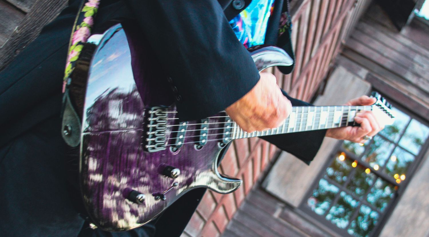 Musician playing a purple electric guitar with a decorative strap outdoors on a wooden deck.