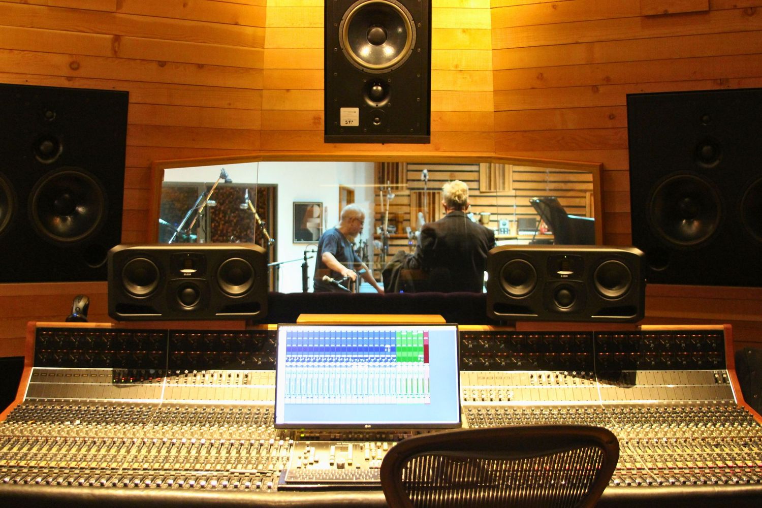 Professional recording studio with mixing console, laptop, monitors, and engineers visible through glass booth.