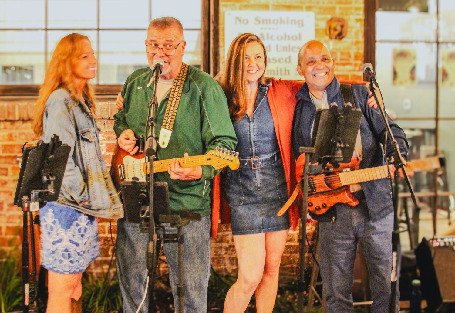 Four musicians smile on stage, two holding guitars, performing live at an outdoor venue in casual attire.