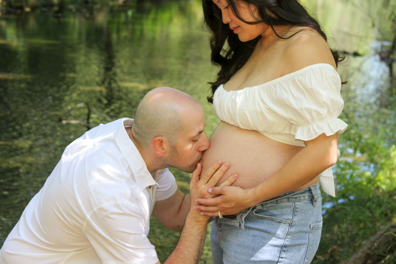 Bald man kissing pregnant woman's belly outdoors near a pond during a maternity photoshoot.
