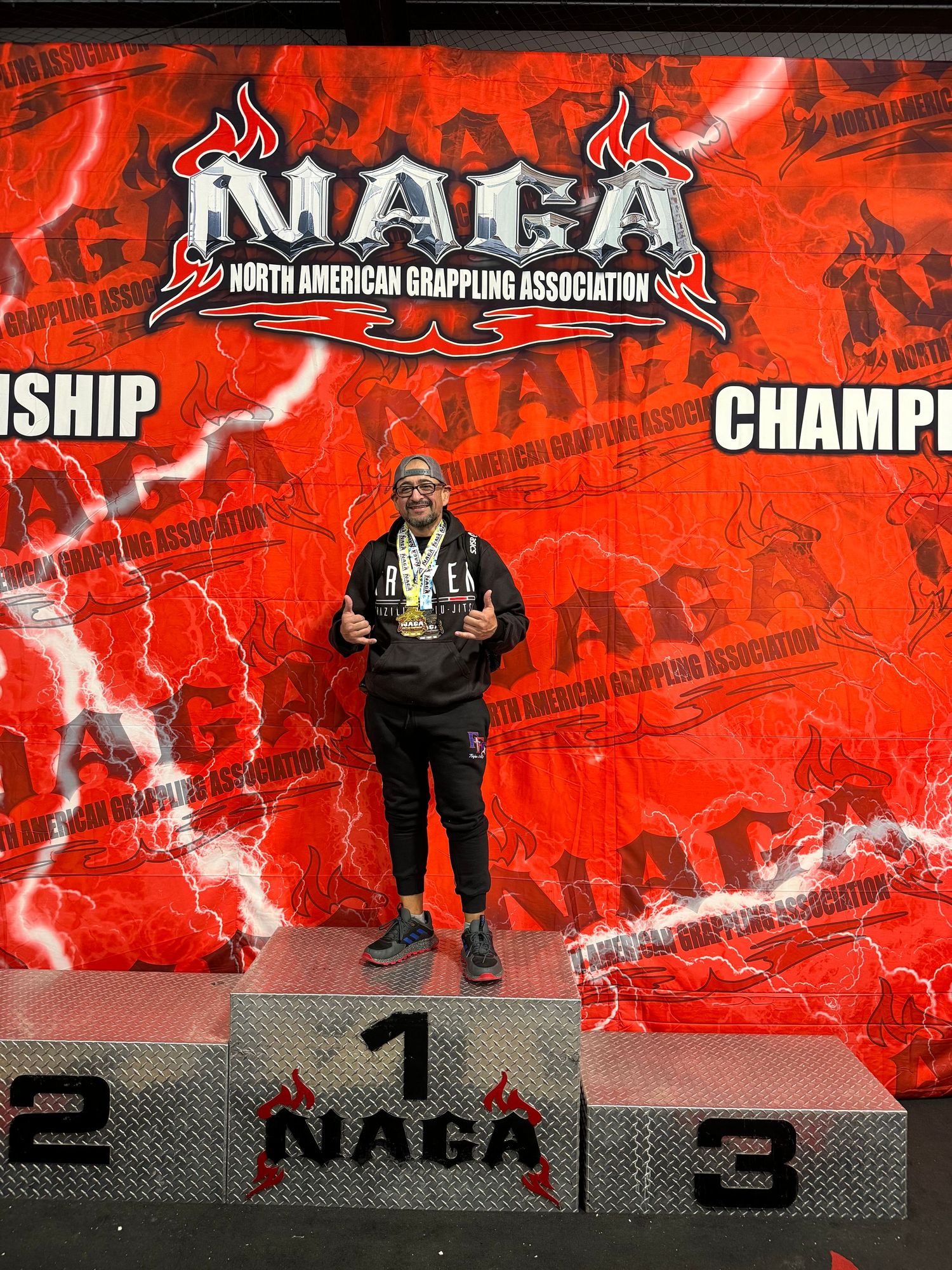 Athlete stands on first place podium at NAGA North American Grappling Association Championship, wearing medal and black outfit.