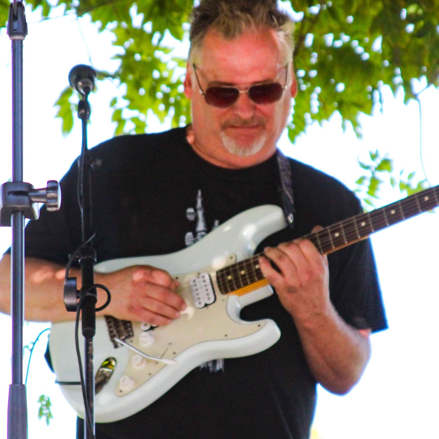 Middle-aged man with sunglasses playing a white Fender Stratocaster guitar outdoors at a live music event.