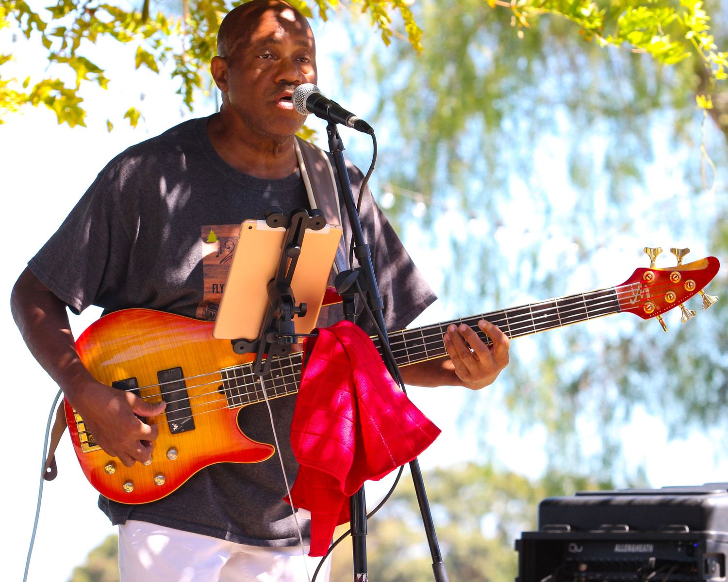A musician plays a sunburst bass guitar and sings into a microphone at an outdoor concert.
