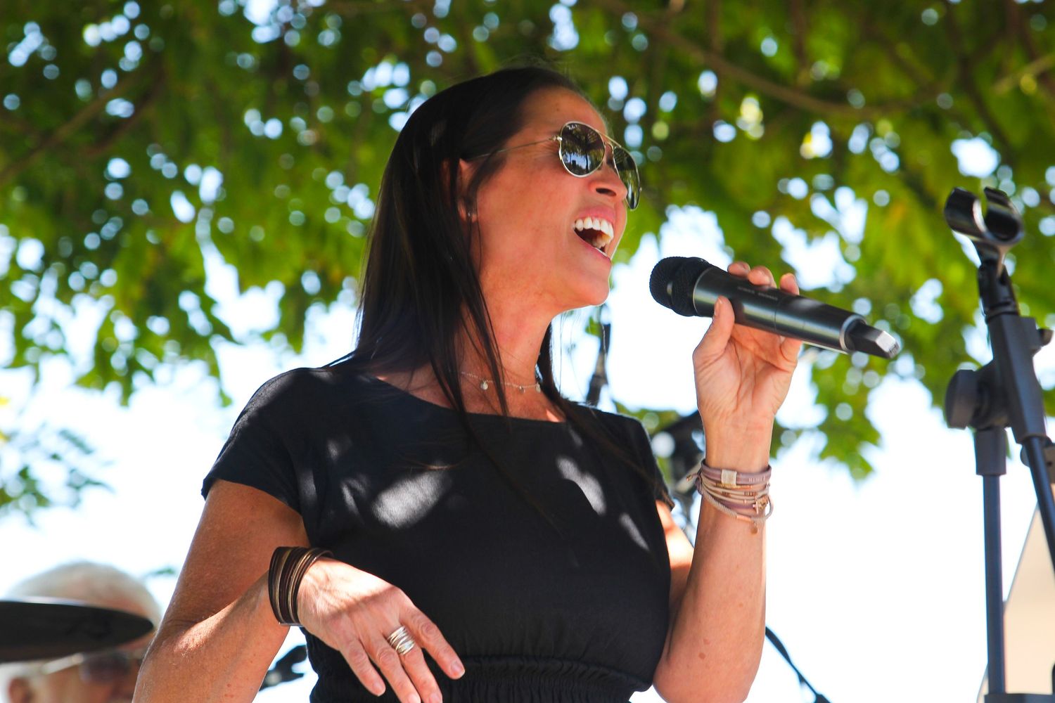 Woman in black dress singing into microphone outdoors at live music event under green trees.