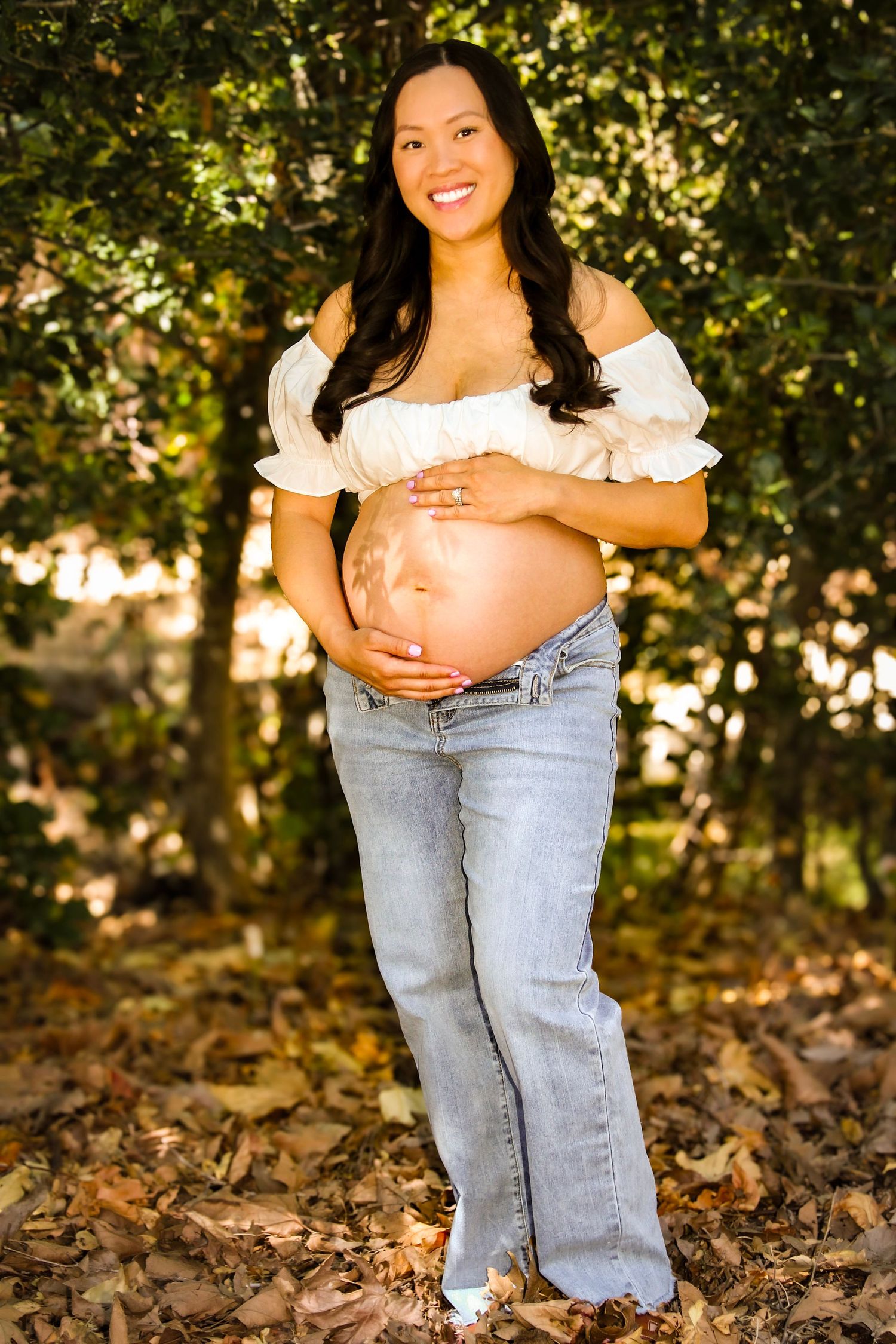 Smiling pregnant woman in white off-shoulder top and jeans cradling her baby bump outdoors among autumn leaves.